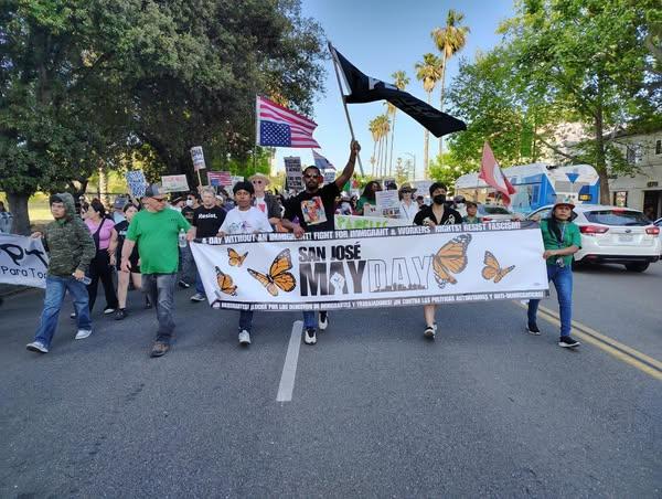 Community members marching with flags and signs, behind a banner that reads “San Jose May Day”