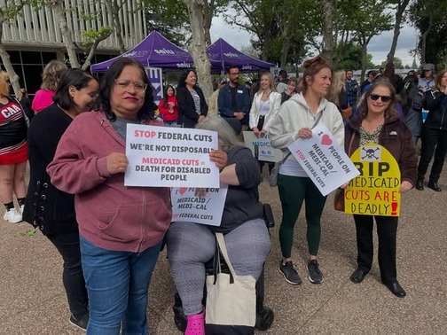 Several community members standing at a rally with three signs that read: “Stop federal cuts. We’re not disposable. Medicaid cuts = death for disabled people” and “I [heart] people on medicaid/medical. Stop federal cuts” and “RIP Medicaid cuts are deadly.”